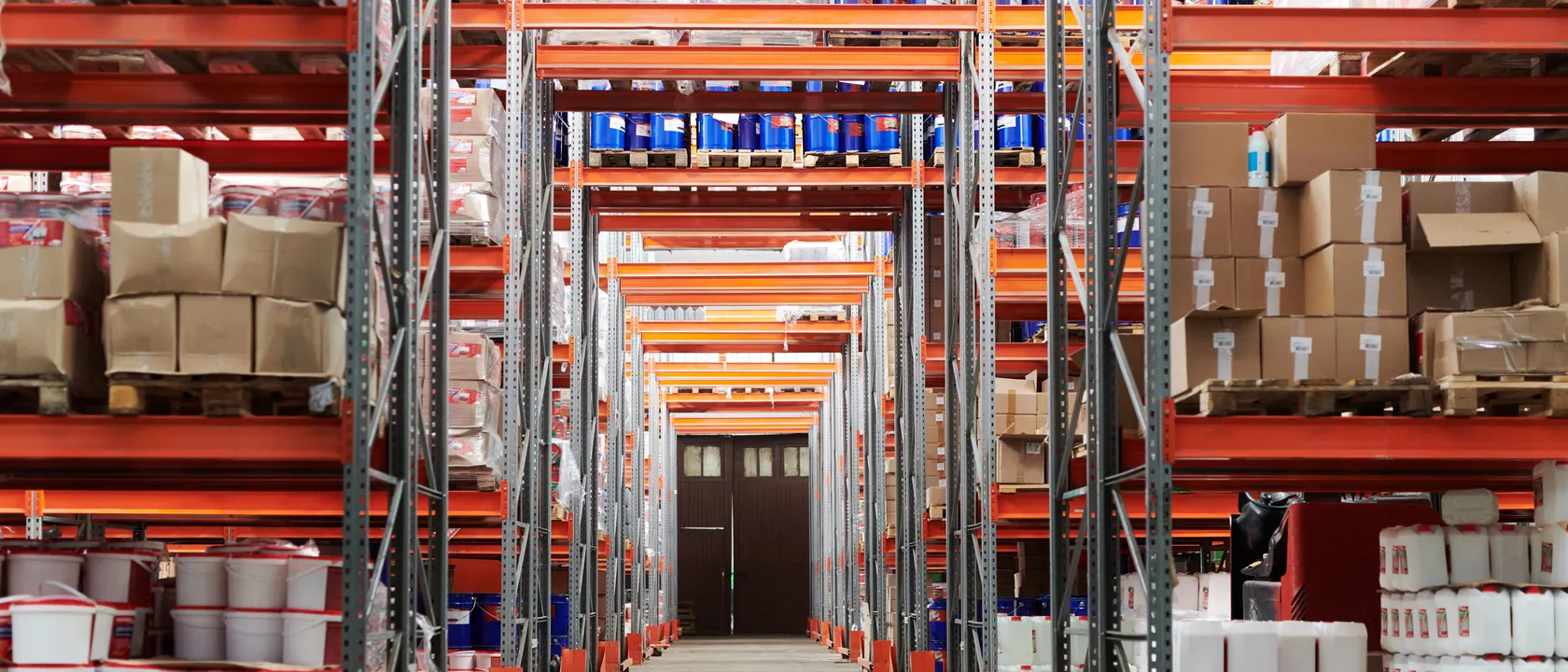A warehouse aisle running between tall pallet racking stacked with boxes and drums
